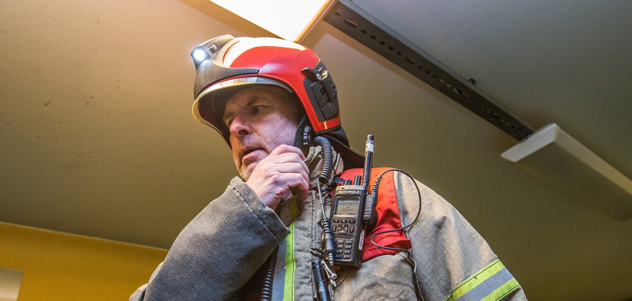 Nærbilde av en brannmann med radioterminal på brystet. Han befinner seg innendørs. 